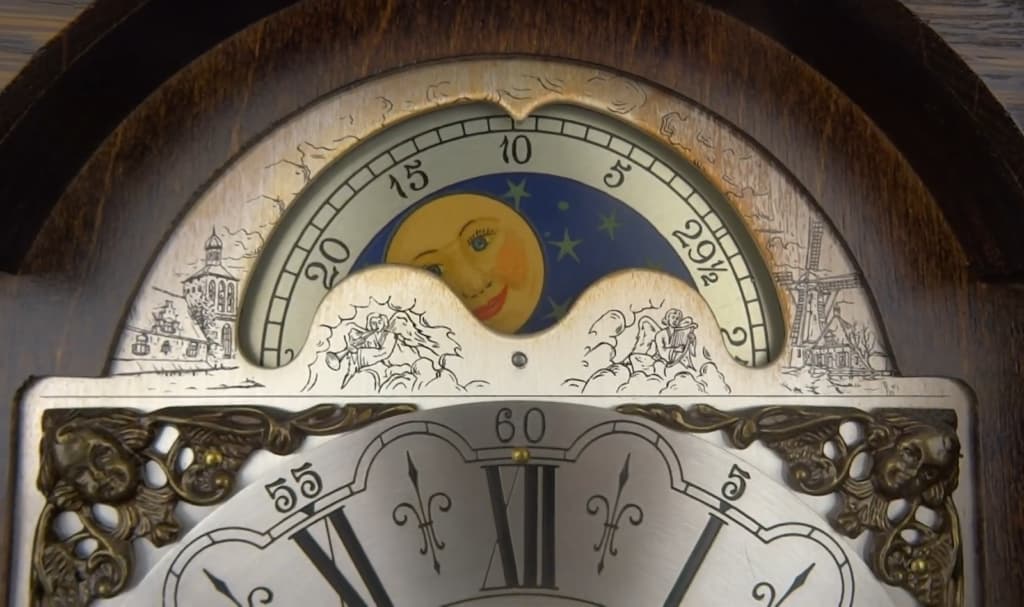 A vintage clock displays a painted moon with a face, with small illustrations of a forest landscape and a map of Europe and Asia